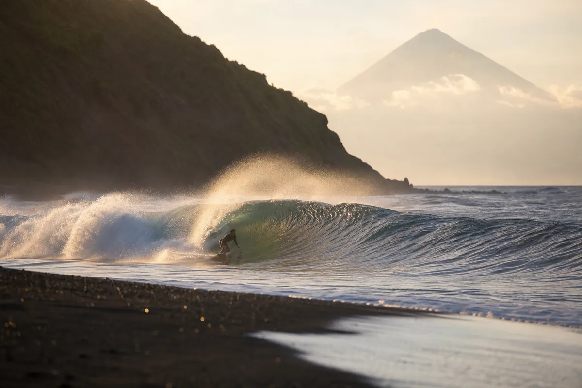 A surfer riding a long left-hand wave along Raglan's black-sand coast with a volcanic cone in the distance